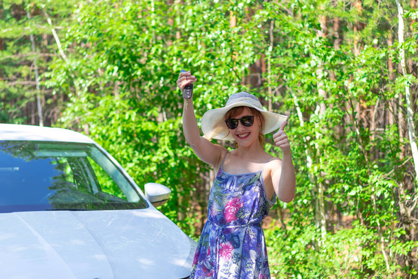 Красивая радостная женщина перед белой машиной с ключом в руке в летний солнечный день на фоне зеленых деревьев. Селективный фокус - Фото, изображение