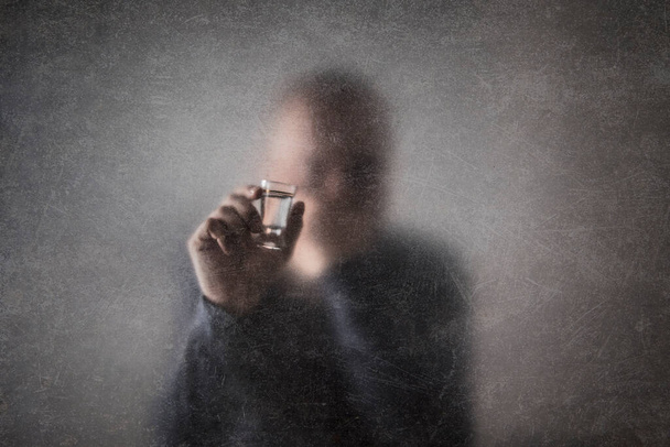 Mann mit einem Glas Wodka hinter einem staubig zerkratzten Glas. Alkoholismus-Konzept. - Foto, Bild