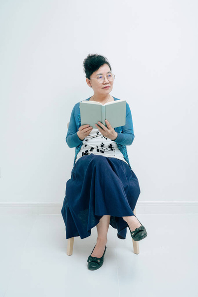 Жінка середнього віку читає книгу, сидячи перед білим тлом
 - Фото, зображення