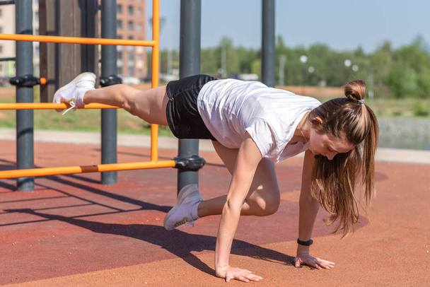 Young athletic woman in a white T-shirt,black shorts and white sneakers going in for sports,.doing an exercise in outdoor gym.Sports healthy lifestyle.Outdoor training. - Fotografie, Obrázek