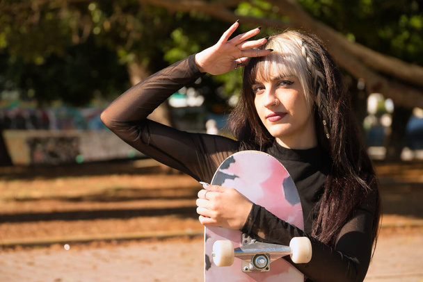 Porträt eines jungen und schönen Mädchens mit Heterochromie und Punkstil, das die Sonne mit der Hand bedeckt, auf einem Bordstein sitzt und ihr Skateboard umarmt. - Foto, Bild