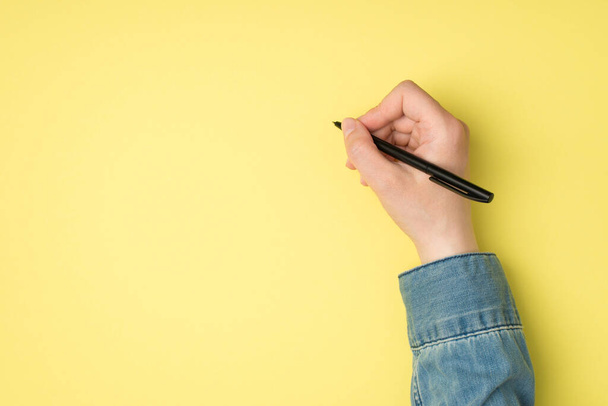 First person top view photo of woman's hand holding black pen on isolated yellow background with blank space - Fotoğraf, Görsel