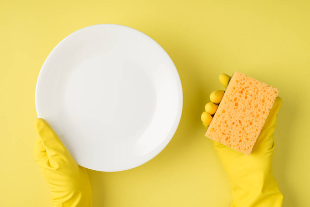 Top view photo of hands in yellow rubber gloves holding white clean plate and yellow sponge on isolated yellow background with copyspace - Фото, зображення