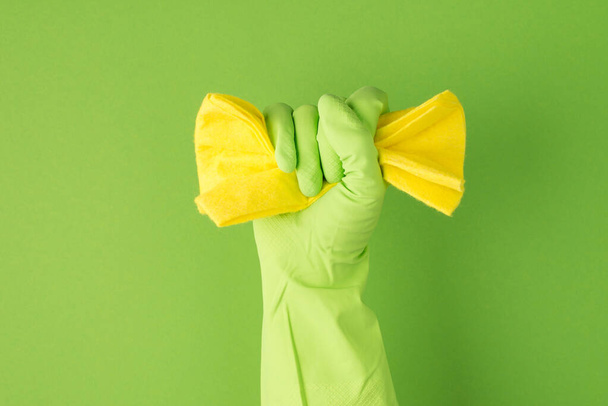 Φωτογραφία του έθεσε το χέρι σε πράσινο γάντι καουτσούκ σφίγγοντας κίτρινη πετσέτα σε απομονωμένο πράσινο φόντο με copyspace - Φωτογραφία, εικόνα