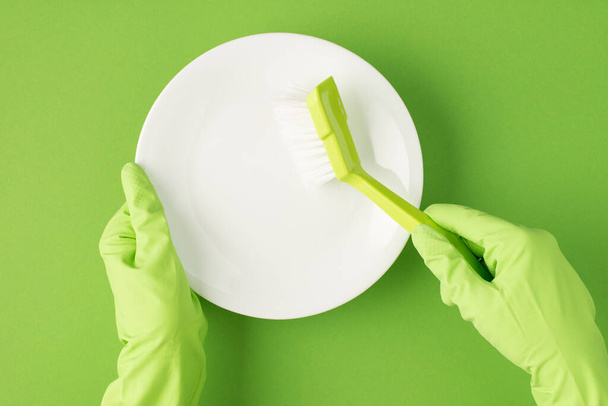 Top view photo of hands in green rubber gloves holding white clean plate and green brush on isolated green background - Photo, Image