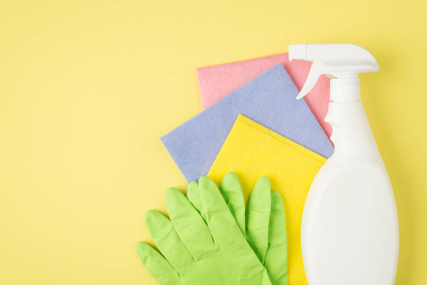 Foto vista superior del detergente en aerosol botella blanca sin etiqueta tres trapos multicolores y guantes de goma sobre fondo amarillo aislado con espacio en blanco - Foto, Imagen
