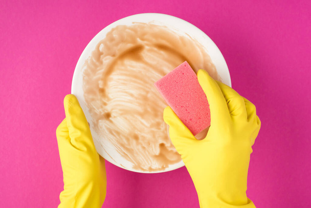 Top view photo of hands in yellow rubber gloves holding dirty plate and using pink scouring pad on isolated pink background - Photo, Image