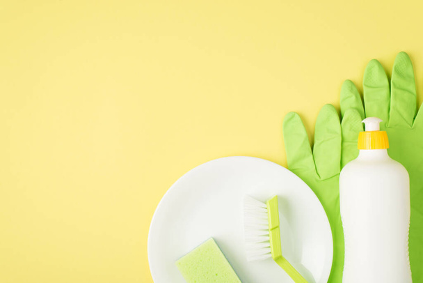 Top view photo of detergent foam bottle without label green sponge and brush on dish rubber gloves on isolated yellow background with copyspace - Photo, Image