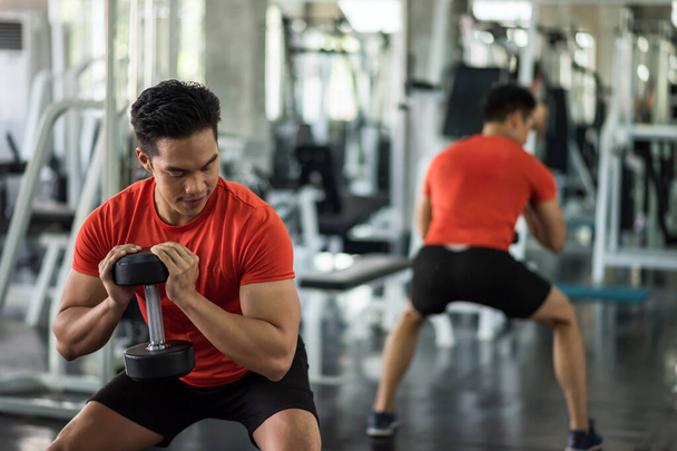 Ázsiai izmos erős fiatalember gyakorolni bicepsz kezek és lábak nehéz súlyzó fitnesz tornaterem sport gyakorló berendezések és a tükörreflexió. Testfelépítés és egészséges életmód koncepció. - Fotó, kép