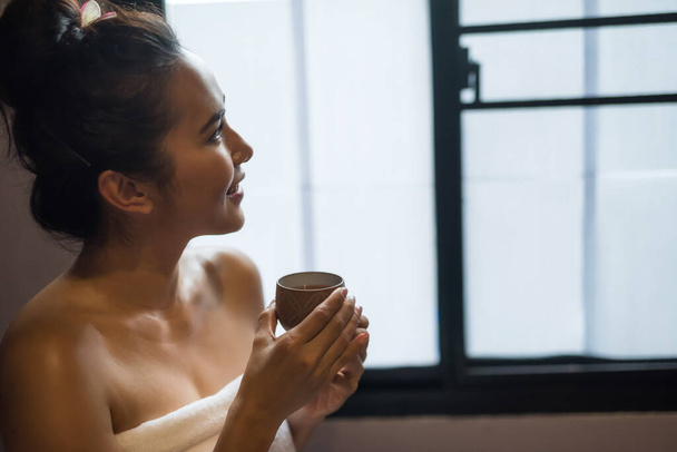 Souriant belle jeune femme asiatique en blanc tenir tasse de thé chaud après avoir eu massage thaï dans le salon de spa. Fille heureuse avec thérapie médicale alternative. - Photo, image
