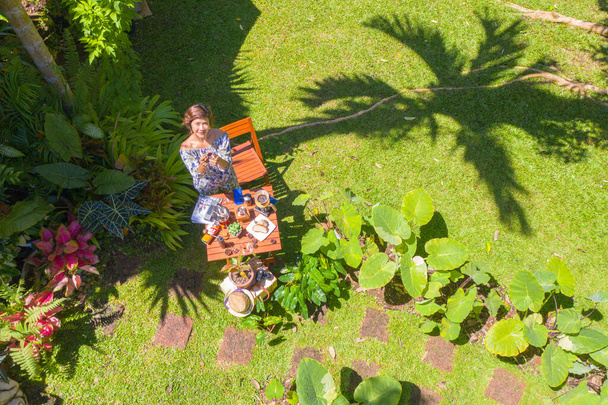 Жінка тримає склянку гарячої кави в зеленому саду. Жінка відпочиває в зеленому саду з кавою, встановленою на дерев'яному столі. зелений фон саду
. - Фото, зображення