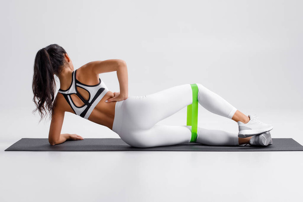 Fitness žena dělá clamshell cvičení pro hýždě s páskou odporu na šedém pozadí. Sportovní dívka cvičení ven - Fotografie, Obrázek