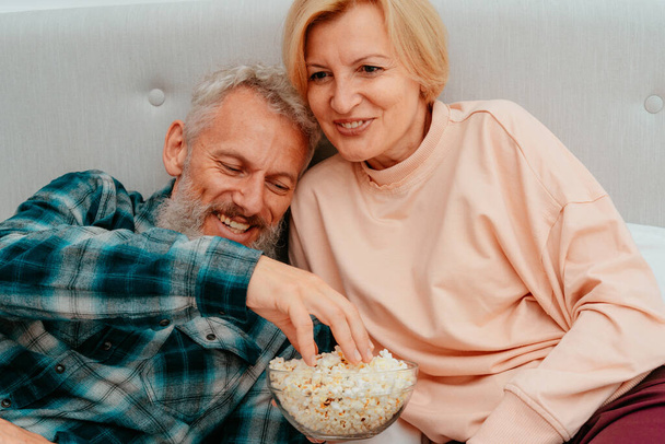 Husband and wife watch a film on the bed and eat popcorn - Photo, Image