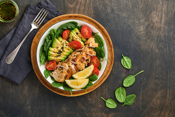 Fresh green salad with grilled chicken fillet, spinach, tomatoes, avocado, lemon and black sesame seeds in white bowl on old wooden dark table background. Nutrition Diet Concept. Top view . - Φωτογραφία, εικόνα