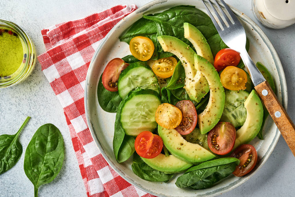Avocado, red, yellow, black cherry tomato, spinach and cucumber fresh salad with spices pepper and olive oil in grey bowl on grey slate, stone or concrete background. Healthy food concept. Top view. - Foto, Bild