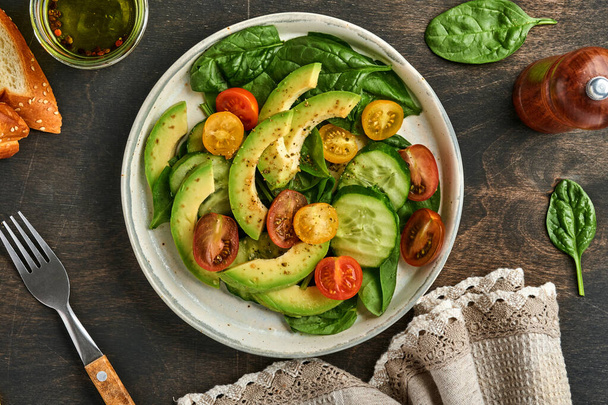 Avocado, red, yellow, black cherry tomato, spinach and cucumber fresh salad with spices pepper and olive oil in grey bowl old wooden table background. Healthy food concept. Top view. - Фото, изображение