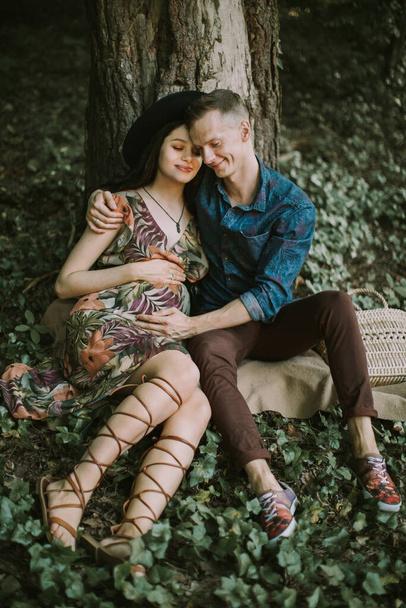Pregnant couple, sitting under the tree in the beautiful forest, enjoying their walk together. - Foto, afbeelding