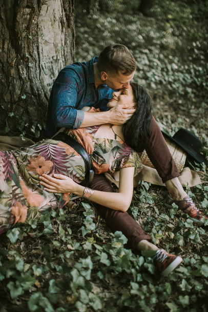 man, kissing her pregnant wife in forehead, while sitting under the tree in the forest outdoors - Foto, Imagem