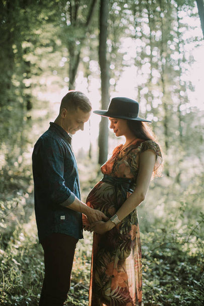 man and his pretty pregnant wife, standing on the background of beautiful sunset in summer forest - Foto, imagen
