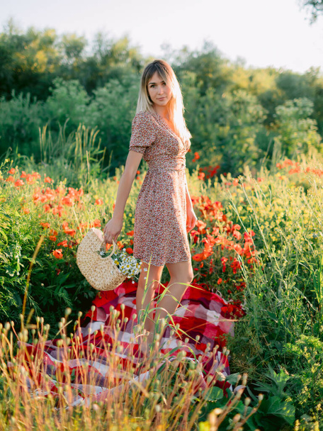 Blonde woman in outdoor with poppy flowers at sunset. Woman in summer dress and wicker handbag with flowers - Φωτογραφία, εικόνα