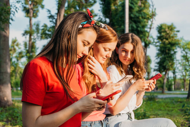 Tres amigas viendo teléfonos inteligentes móviles y divirtiéndose sentadas en el banco en el parque público. Gen z, La adicción a los dispositivos para adolescentes, Desintoxicación digital, cultura juvenil, tecnología, amistad social - Foto, Imagen