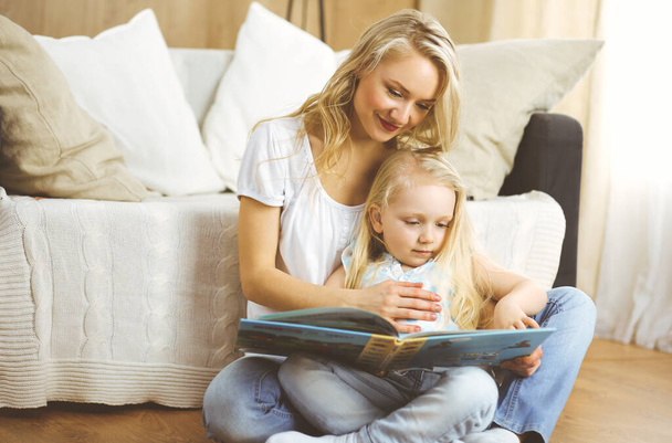 Happy family. Blonde young mother reading a book to her cute daughter while sitting at wooden floor. Motherhood concept - Zdjęcie, obraz