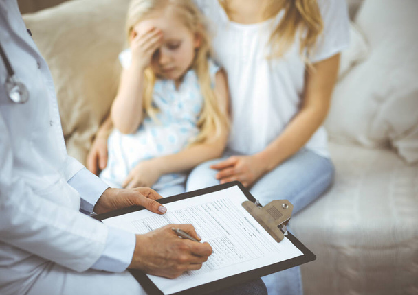 Doctor and patient. Pediatrician using clipboard while examining little girl with her mother at home. Sick and unhappy child at medical exam. Medicine concept - Foto, afbeelding