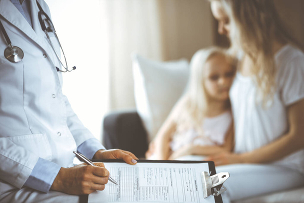 Doctor and patient. Pediatrician using clipboard while examining little girl with her mother at home. Sick and unhappy child at medical exam. Medicine concept - Φωτογραφία, εικόνα
