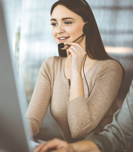 Two young people in headsets are talking to the clients, while sitting at the desk in an office. Focus on woman. Call center operators at work - Photo, Image