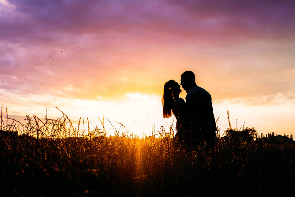 ロマンチックな休暇、ハネムーンへのロマンチックな目的地、ソロ旅行。自然夕日黄色と紫の空の背景に愛のシルエットのカップル。牧草地で夕日を楽しむ若いカップル. - 写真・画像