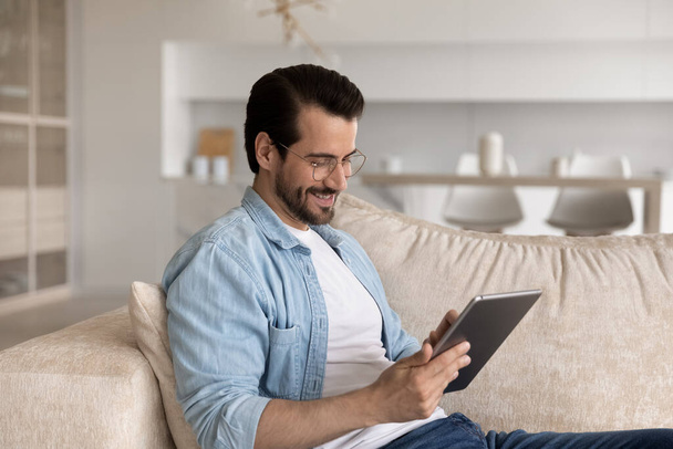 Smiling man in glasses using tablet, sitting on cozy couch - Фото, зображення