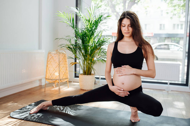 Patient pregnant woman sitting in a side lunge, practicing yoga alone. She's wearing black top and pants. She's holding her hand on her belly. Cars driving in the background. - Фото, изображение