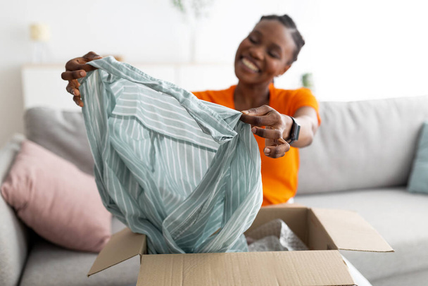 Zufriedene Käufer. Lächelnde schwarze Frau entpackt Paket nach der Lieferung, hält Bluse in der Hand, sitzt zu Hause auf Couch - Foto, Bild