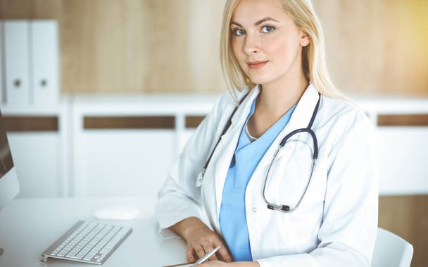Woman-doctor at work while sitting at the desk in sunny clinic. Blonde cheerful physician filling up medication history record form - Фото, изображение