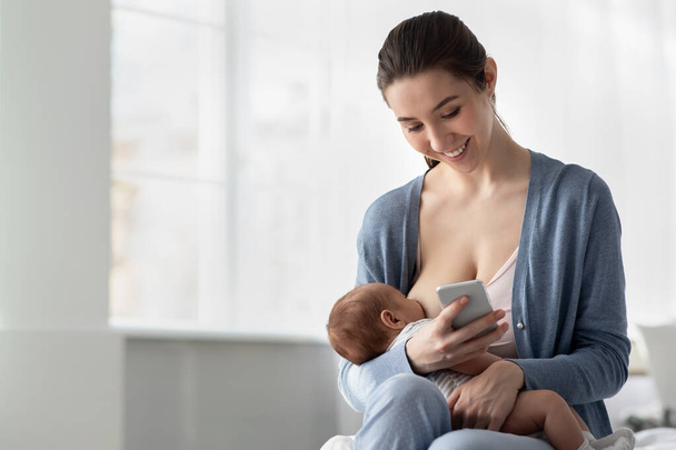Onnellinen nuori äiti käyttää älypuhelinta imettäessään vastasyntynyttä vauvaansa - Valokuva, kuva