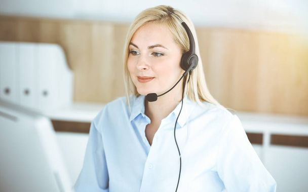 Blonde business woman sitting and communicated by headset in call center in sunny office. Concept of telesales business - Fotoğraf, Görsel