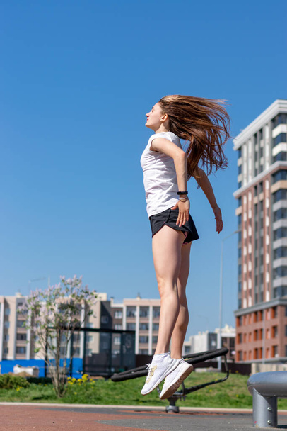 Positive young athletic woman in a white T-shirt,black shorts and white sneakers is jumping.Sports healthy lifestyle.Outdoor training.Urban landscape in the background.Copy space for text. - Foto, immagini