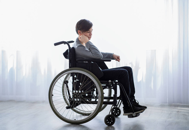 Напряженный мальчик-инвалид в инвалидной коляске чувствует себя одиноким и расстроенным, сидя у окна дома - Фото, изображение