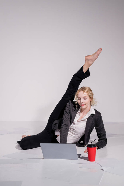 Barefoot ballerina dressed in a business suit poses for a laptop and drinks coffee. Flexible woman works at the computer - Φωτογραφία, εικόνα