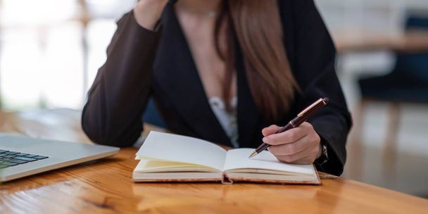 Закройте руки женщины ноутбуком, ноутбуком и ручкой, делая заметки в деловом кабинете - Фото, изображение