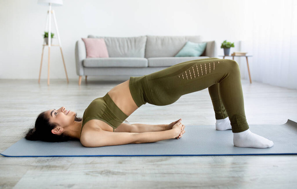 Seitenansicht einer Inderin bei Bauchmuskelübungen, die während ihres häuslichen Trainings in Halb-Brücke-Yoga-Pose steht - Foto, Bild