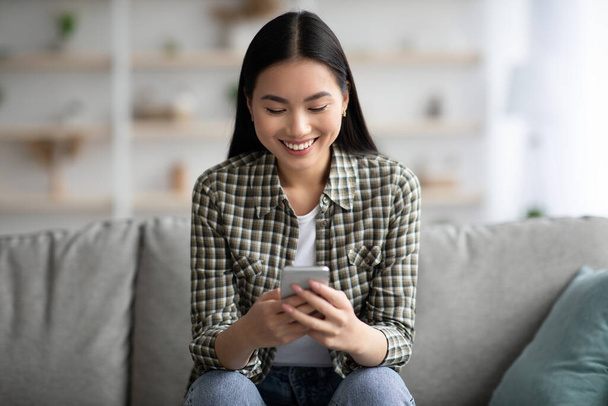 Happy aziatische vrouw zitten op de bank met smartphone, chatten - Foto, afbeelding