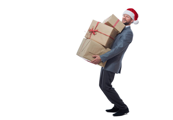 Handsome young man in a suit wearing Christmas hat holding a pile of presents - Фото, изображение