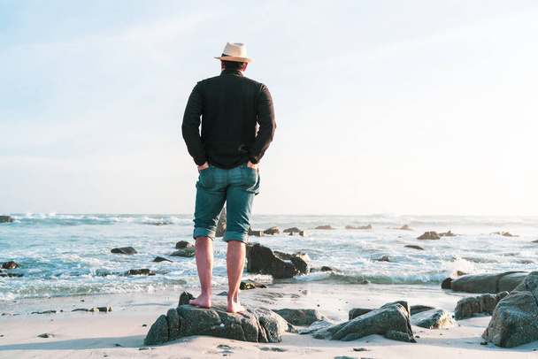 un giovane uomo in piedi sulla roccia spiaggia al tramonto fissando l'orizzonte al tramonto - Foto, immagini