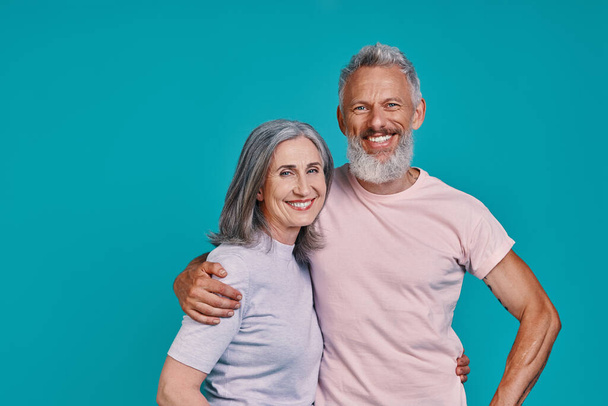 Happy senior couple looking at camera and smiling while standing together against blue background - Фото, изображение