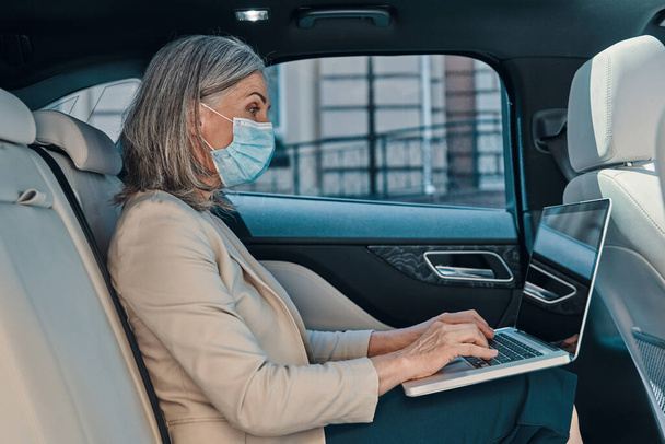 Reife schöne Frau in schützender Gesichtsmaske mit Laptop, während sie auf dem Rücksitz im Auto sitzt - Foto, Bild