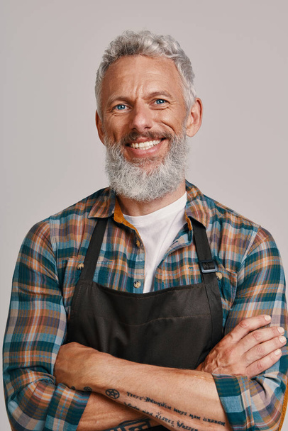 Senior man in apron smiling and looking at camera while standing against gray background - Фото, изображение