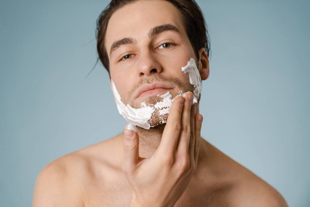 Portrait of a mid aged brunette shirtless man applying shaving foam on his face over blue wall background - Valokuva, kuva