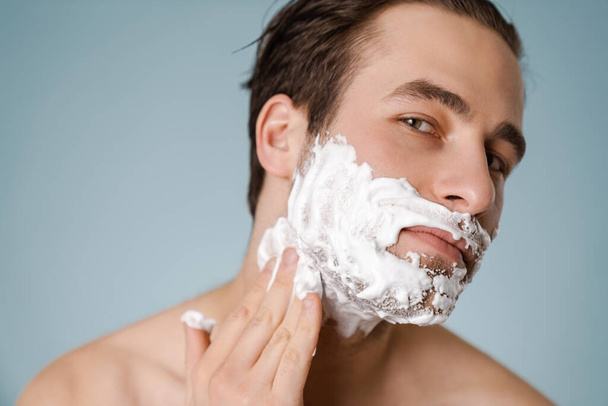 Portrait of a mid aged brunette shirtless man applying shaving foam on his face over blue wall background - Foto, afbeelding