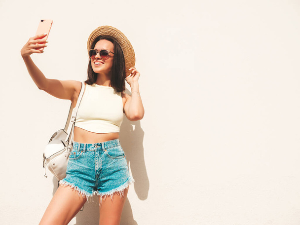 Portrait of young beautiful smiling hipster woman in trendy summer jeans shorts and hat. Sexy carefree model posing in the street near white wall.Positive model outdoors with briefcase. Taking selfie - Фото, изображение